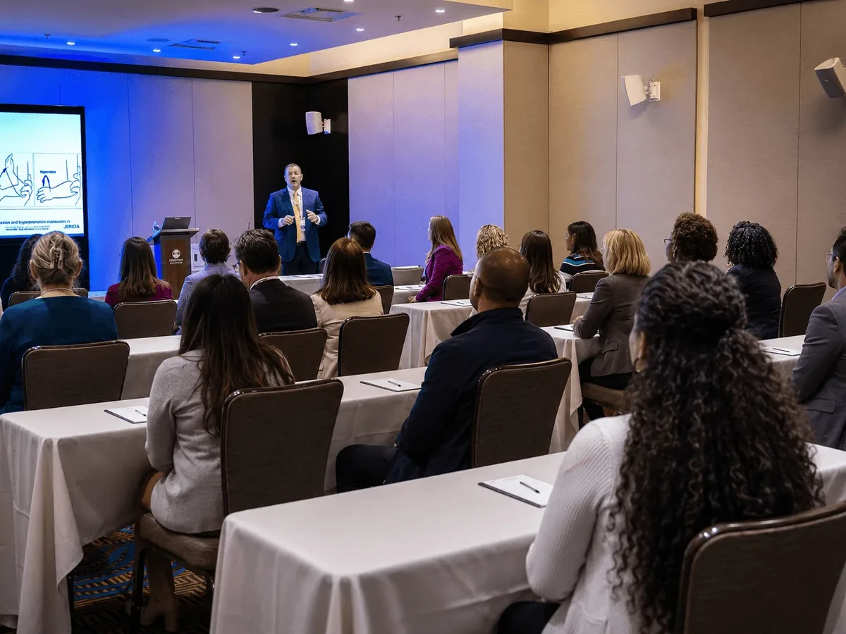 A speaker presenting a clinical session to attendees seated at classroom-style tables.
