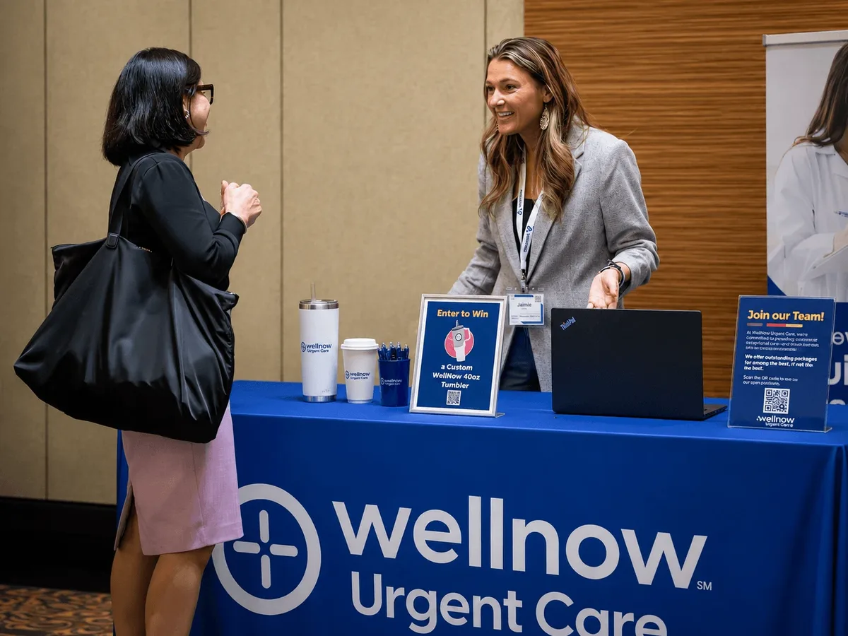 An exhibitor talking with an attendee across a branded urgent care booth in the exhibit hall.