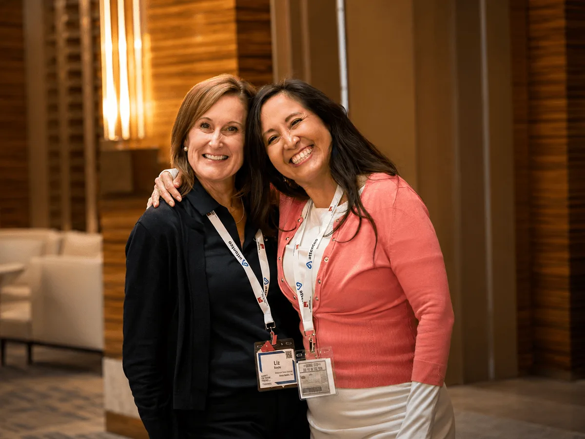 Two conference attendees in lanyards smiling together in the hotel lobby between sessions.